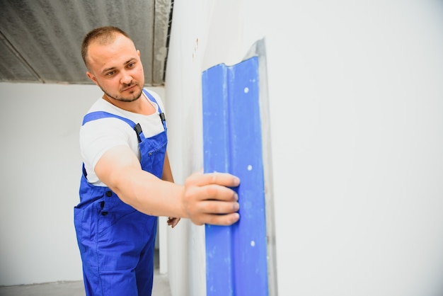 De bouwer corrigeert zorgvuldig de onregelmatigheden van de muur met een troffel. Bouwer in werkkleding tegen een grijze muur. Foto stukadoor aan het werk.