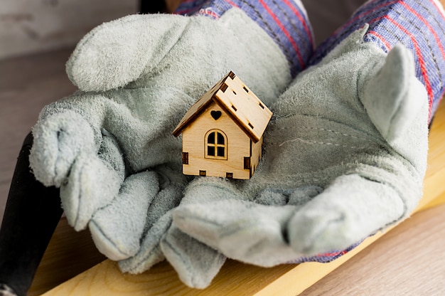 De bouwer houdt in werkhandschoenen model van het huis tegen de achtergrond van bouwmaterialen en hulpmiddelen.