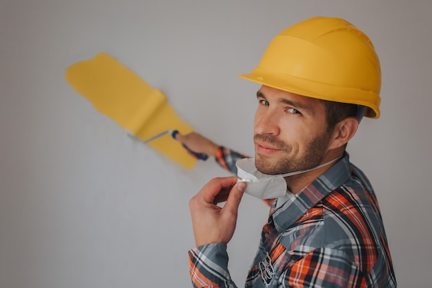 De bouwer werkt op de bouwplaats en meet het plafond. Een arbeider in een oranje helm en een verfroller schildert de muur.