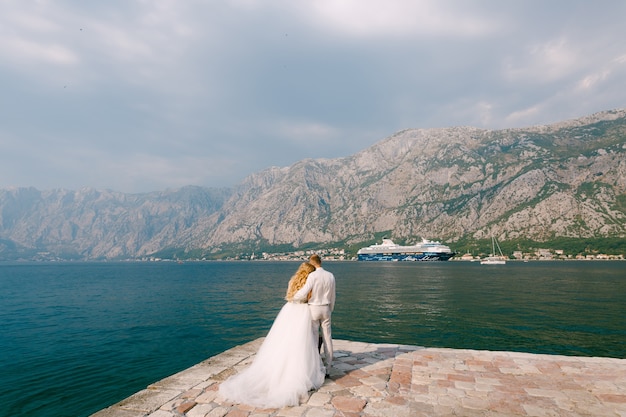 De bruid en bruidegom omhelzen elkaar op de pier in de baai van Kotor, een cruiseschip vaart voorop