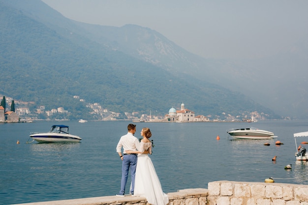 De bruid en bruidegom omhelzen elkaar op de pier in de buurt van het oude centrum van Perast boten die in de buurt drijven