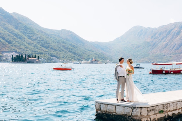 de bruid en bruidegom staan naast elkaar op de pier in de baai van de Kotor-eilanden van Perast