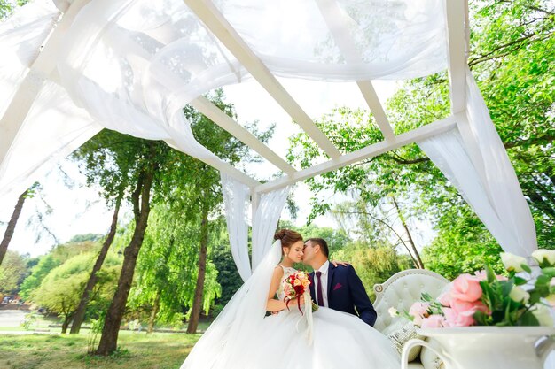 De bruid en bruidegom zitten op een witte bank, knuffelend onder een boog gemaakt van vliegende stof.