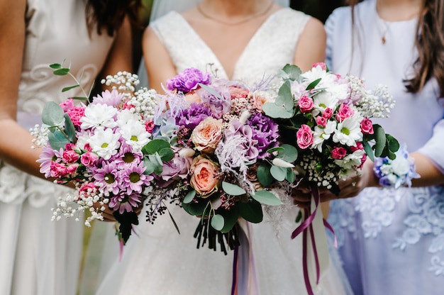 De bruid en bruidsmeisjes in een elegante jurk staan en houden handboeketten van pastelroze bloemen en groen met lint vast.