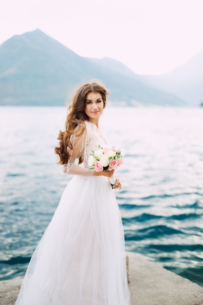 De bruid heeft een boeket rozen in haar handen en staat op de pier in de baai van Kotor