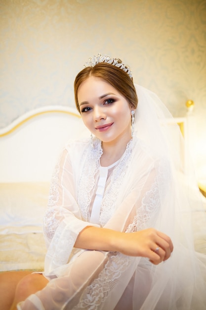 De bruid in een witte jas op het bed op een huwelijksochtend