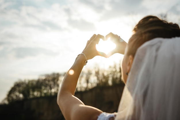 De bruid kijkt naar de zon door haar gesloten handpalmen in de vorm van een hart