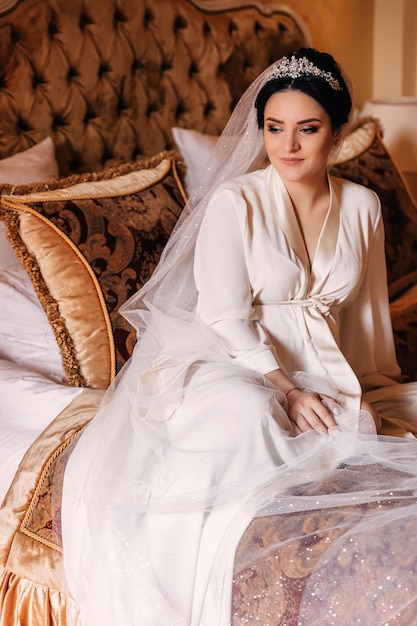 De bruid zit op een elegant bed tussen de kussens. meisje in een wit gewaad en bruidssluier met tiara.