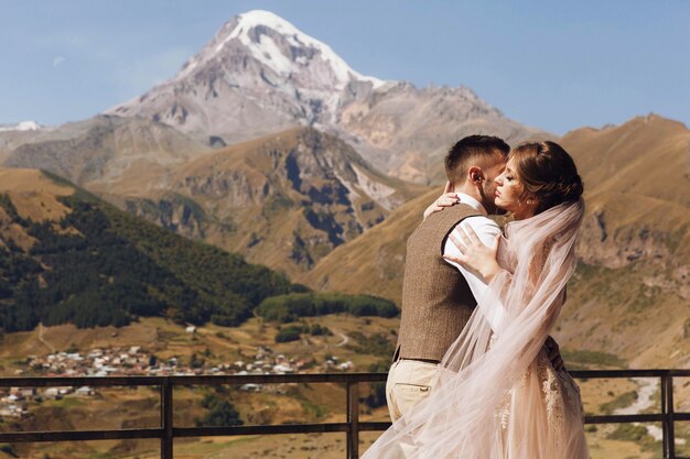 De bruidegom in moderne kostuum en de bruid in charmante roze kleding stellen op de terasse met grote bergmening in Georgië