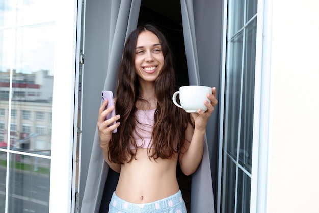 De brunette drinkt koffie op het balkon en sms't aan de telefoon