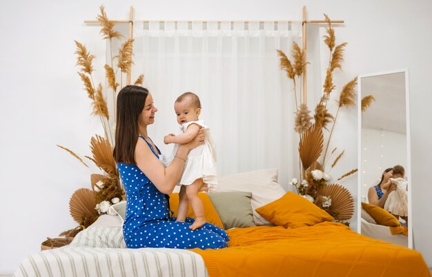 De brunette moeder zit zijwaarts op het bed en speelt met haar dochtertje in een feestelijke jurk