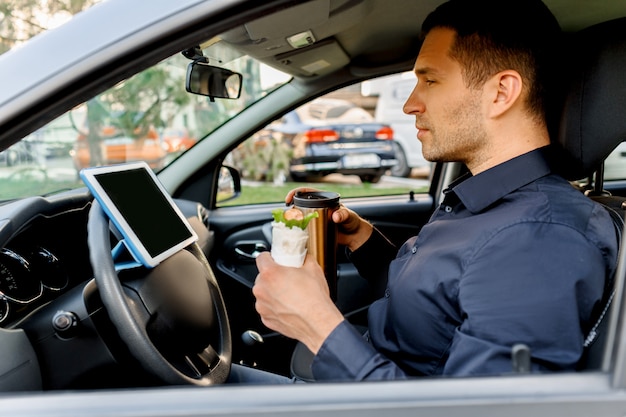De chauffeur kijkt tijdens de lunch naar films of tv-programma's op de tablet. Stoppen voor een hapje. Man eet snack in de auto en drinkt koffie of thee.