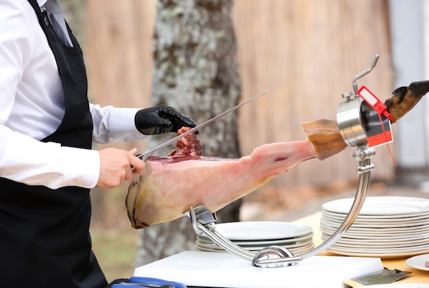 De chef-kok snijdt de ham. afbeelding van een hamsnijder die met een mes een dun plakje ham snijdt. Gedroogde Ham, Serranoham, Acorn, Iberische, Italiaanse of Parma Raw Prosciutto