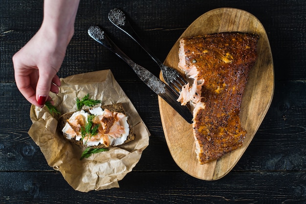 De chef-kok strooit peterselie op een broodje met gerookte zalmfilet op zwart brood en zachte kaas