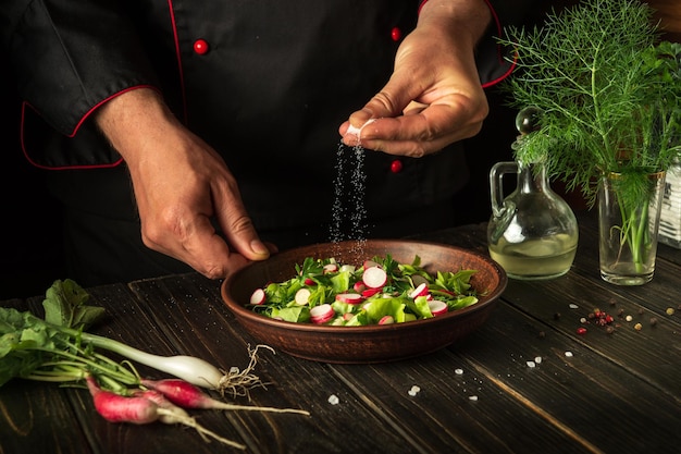 De chef-kok strooit zout op de salade van verse groenten in het bord het bereiden van gezond voedsel in de keuken