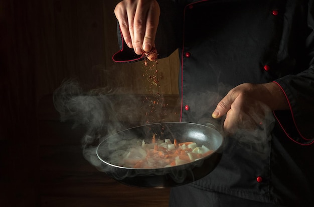 De chef-kok voegt kruiden toe aan gebakken wortelen met uien in een koekenpan