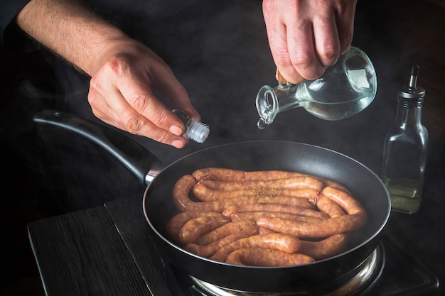 De chef-kok voegt olie toe aan de pan met rauwe vleesworst. Voorbereiding voor het koken van worstjes in de keuken van een restaurant of café