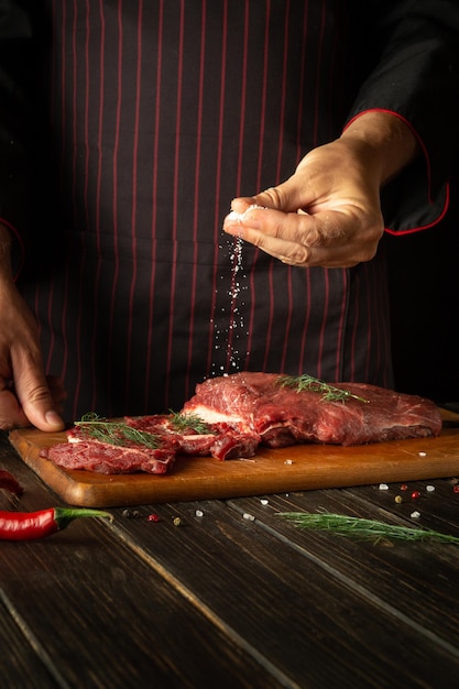 De chef-kok zout rauw vers rundvlees voor het braden of grillen Werkomgeving in de keuken