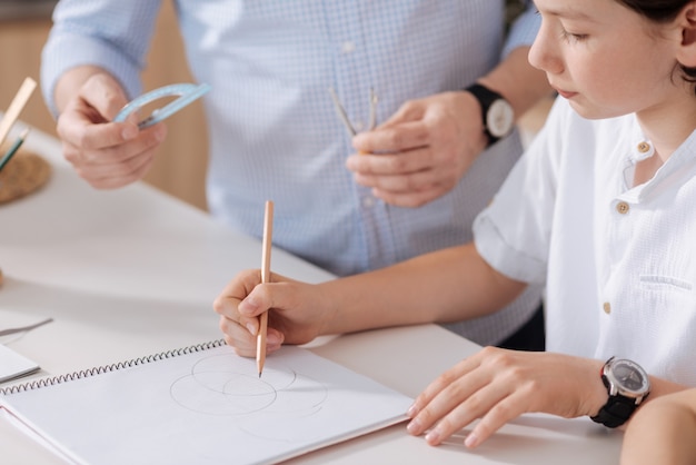 De close-up van een schattige kleine schooljongen die cirkels tekent met een potlood terwijl zijn vader naast hem staat en klaar staat om hem een gradenboog en een kompas te overhandigen