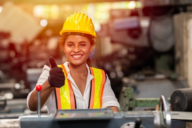 Foto de de tienerarbeider van het meisje met veiligheidshelm toont duim die als arbeid in de industriefabriek werken met staalmachine.