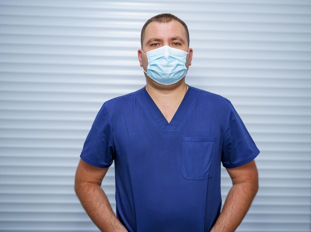 De dokter in blauw uniform met een medisch masker op een lichte achtergrond. Medisch werker op kantoor.