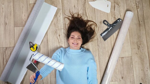 Foto de donkerbruine vrouw met lang los haar die blauwe sweater draagt houdt rol die op vloer ligt. lady glimlacht tussen verspreide bouwmaterialen en gereedschappen op parket bovenaanzicht.