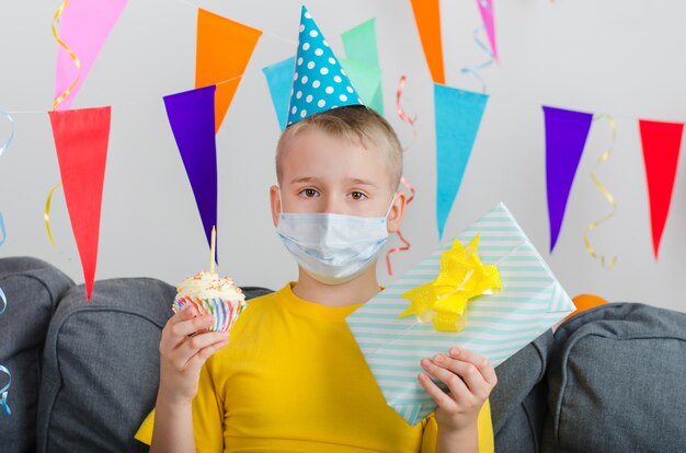 De droevige jongen in het gezichtsmasker van de geneeskunde met in hand giften viert verjaardag