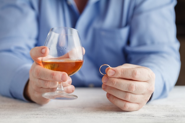 Foto de droevige mens houdt een trouwring dichtbij het glas alcoholische drank terwijl het zitten bij keuken, close-up