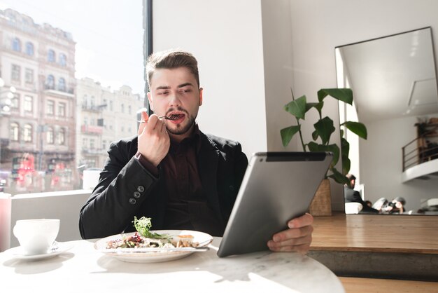 De drukke man zit in een café, dineert en bekijkt video's op de tablet.