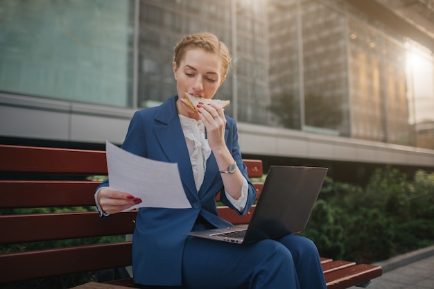 De drukke vrouw heeft haast, ze heeft geen tijd, ze gaat buiten een snack eten. werknemer eten en werken met documenten op de laptop op hetzelfde moment. zakenvrouw meerdere taken uitvoeren. mult.