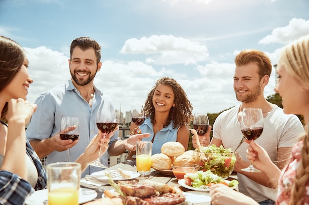 De eerste zomerbarbecuegroep van gelukkige jonge mensen eet vers voedsel, pratend en drinkend