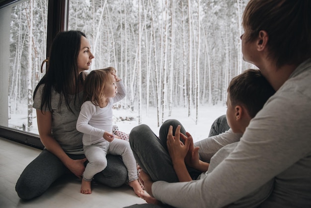 De familie zit bij het raam en kijkt naar het winterbos. Goede nieuwjaarsgeest. Ochtend in pyjama.