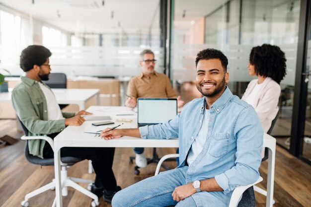 De focus ligt op een glimlachende jonge Arabische professional in een denim shirt die voor een moderne kantoorruimte zit