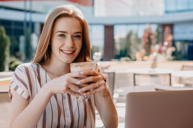 De foto van een charmant roodharig meisje dat haar cocktail drinkt en geniet van haar pauze voordat ze aan de computer gaat werken