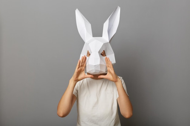De foto van een onbekende vrouw die een wit T-shirt en een papieren konijnenmasker draagt, houdt de handen in de buurt van de mond terwijl ze luid schreeuwt terwijl ze een aankondiging doet en een advertentie vertelt die geïsoleerd staat over een grijze achtergrond