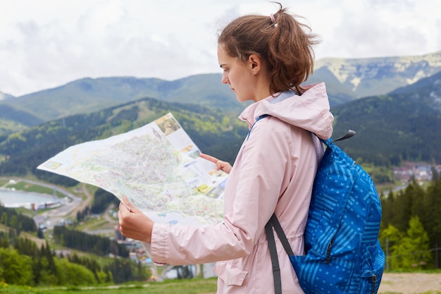 De foto van jonge reizigersvrouw vindt manierrichting met in hand kaart, dame het stellen tegen mooi landschap die bovenop berg zijn