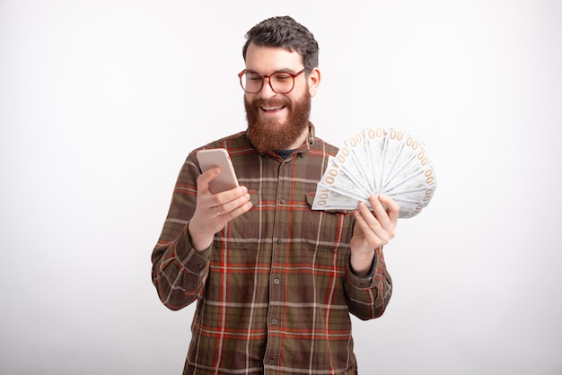 De gebaarde mens die glazen dragen bekijkt zijn telefoon terwijl het houden van een bos van geld.