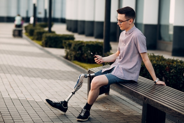 De gehandicapte jonge mens die met voetprothese zitten en houdt mobiele telefoon openlucht