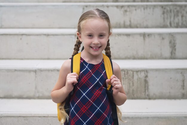 Foto de gele rugzak van het blonde schoolmeisje straalt van vreugde als ze in haar jurk naar school gaat