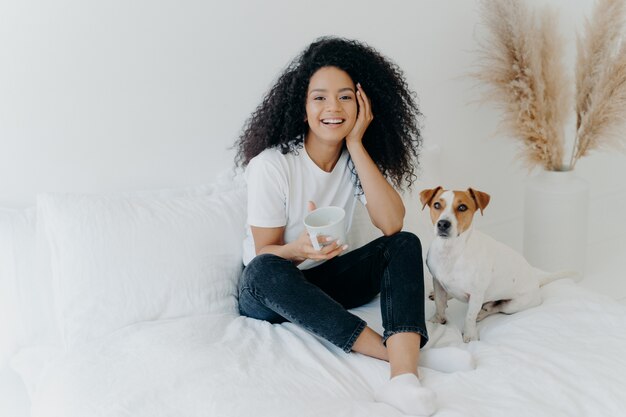 De gelukkige aantrekkelijke etnische vrouw met krullend haar draagt witte t-shirt, jeans en sokken, glimlacht aangenaam, drinkt thee in comfortabel bed, stelt met hond, heeft lui weekend. Mensen, rust, dieren concept