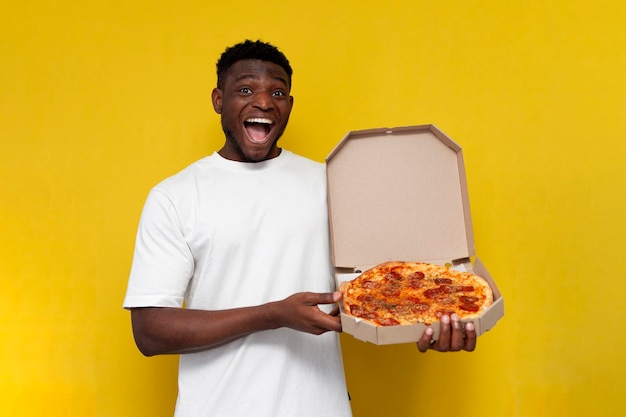 De gelukkige afro-amerikaanse man in een wit t-shirt houdt een doos pizza vast en verheugt zich op gele achtergrond