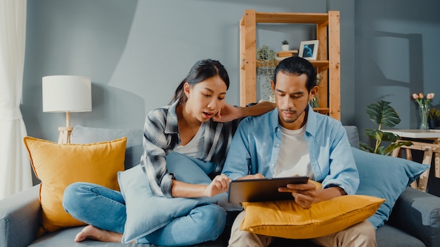 De gelukkige Aziatische jonge aantrekkelijke paarman en vrouw zitten op de tablet van het bankgebruik om online meubilair in nieuw huis te winkelen