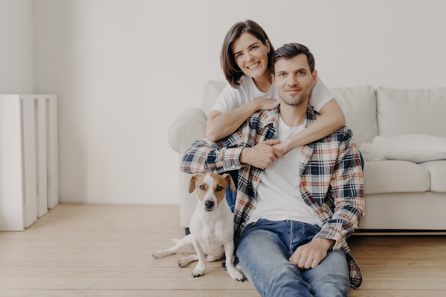 De gelukkige donkerbruine vrouw omhelst echtgenoot met liefde, die in goed humeur is, glimlacht positief. Man, vrouw en hond poseren samen in de woonkamer van een nieuwe woning, genieten van comfort. Verliefde paar binnen