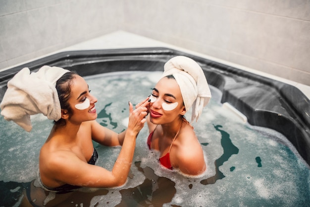 De gelukkige en positieve jonge vrouwen zetten wat ooglap op het gezicht van haar Aziatische vriend. Ze lachen allebei. vrouwen zitten in een hydromassagebad vol met water.