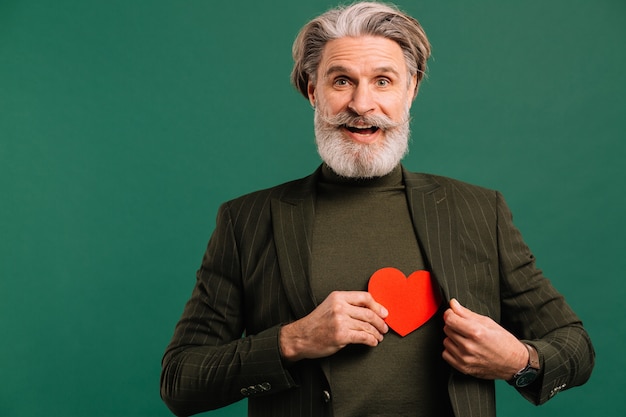 De gelukkige gebaarde en snor rijpe mens trekt een rood hart uit een kaki kostuum die Valentine isoleted groene achtergrond begroeten.