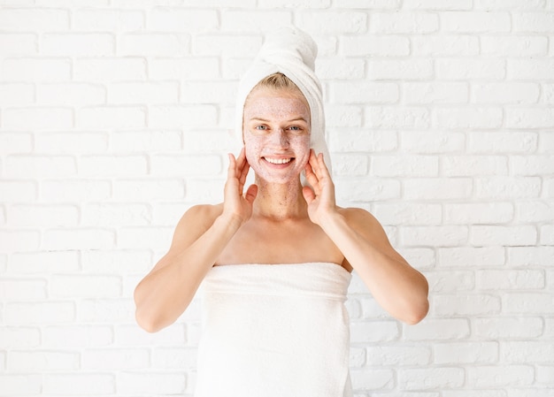 De gelukkige glimlachende jonge vrouw in het witte badhanddoeken van toepassing zijn schrobt op haar gezicht en hals