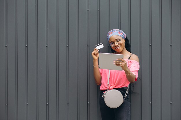 De gelukkige jonge Afrikaanse Amerikaanse vrouw maakt het winkelen in de tablet met creditcard bij de grijze muur