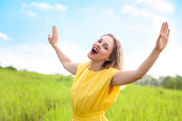 De gelukkige jonge blondevrouw in gele kleding zingt het kijken aan camera op groene weide.