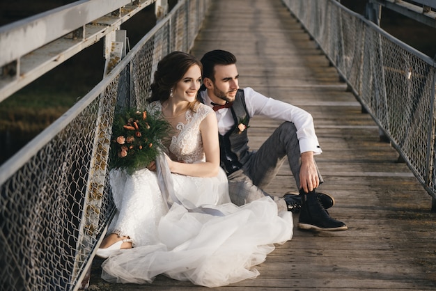 De gelukkige jonge glimlachende bruid en de bruidegom zitten op de hangbrug. zonnige trouwfoto's op een interessante plek