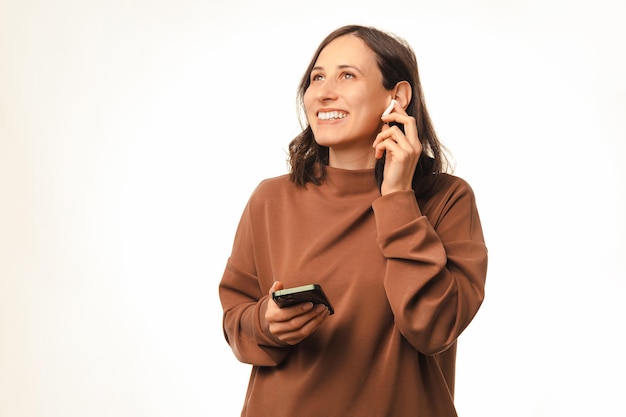 De gelukkige jonge glimlachende vrouw gebruikt haar telefoon en draadloze oortelefoons